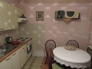 Eine kompakte Küche mit Herd, Spüle und Mikrowelle. Ein Esstisch mit Stühlen und ein kleines gerahmtes Bild an der Wand sind ebenfalls vorhanden.