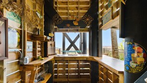 A rustic wooden bar area with a wooden ceiling, shelves with various items, and a view through a large window.