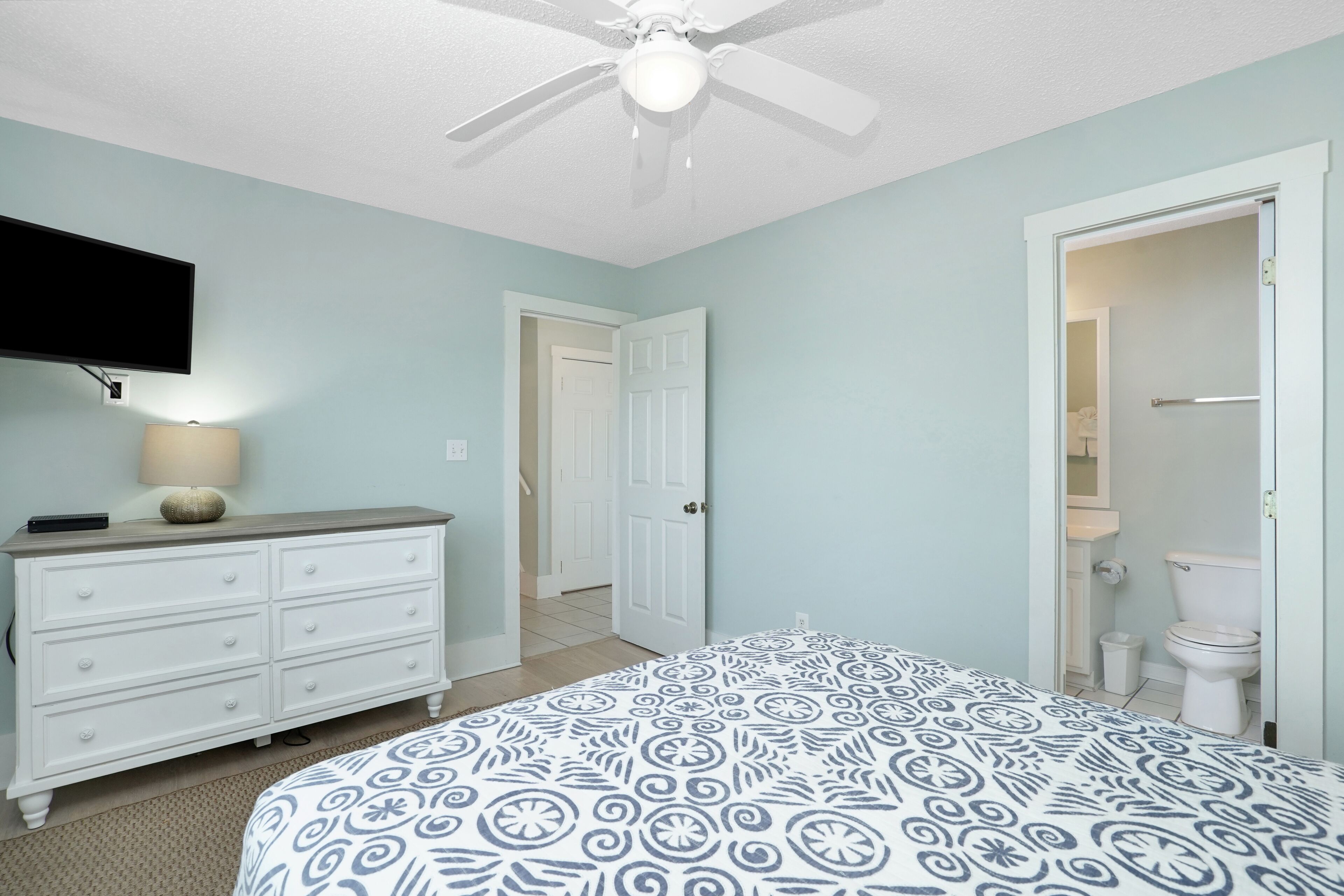 A bedroom with a bed, a dresser, a ceiling fan, and a television mounted on the wall.