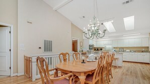 Uma sala de jantar espaçosa com uma grande mesa de madeira rodeada por cadeiras, um candelabro e vista para a cozinha.