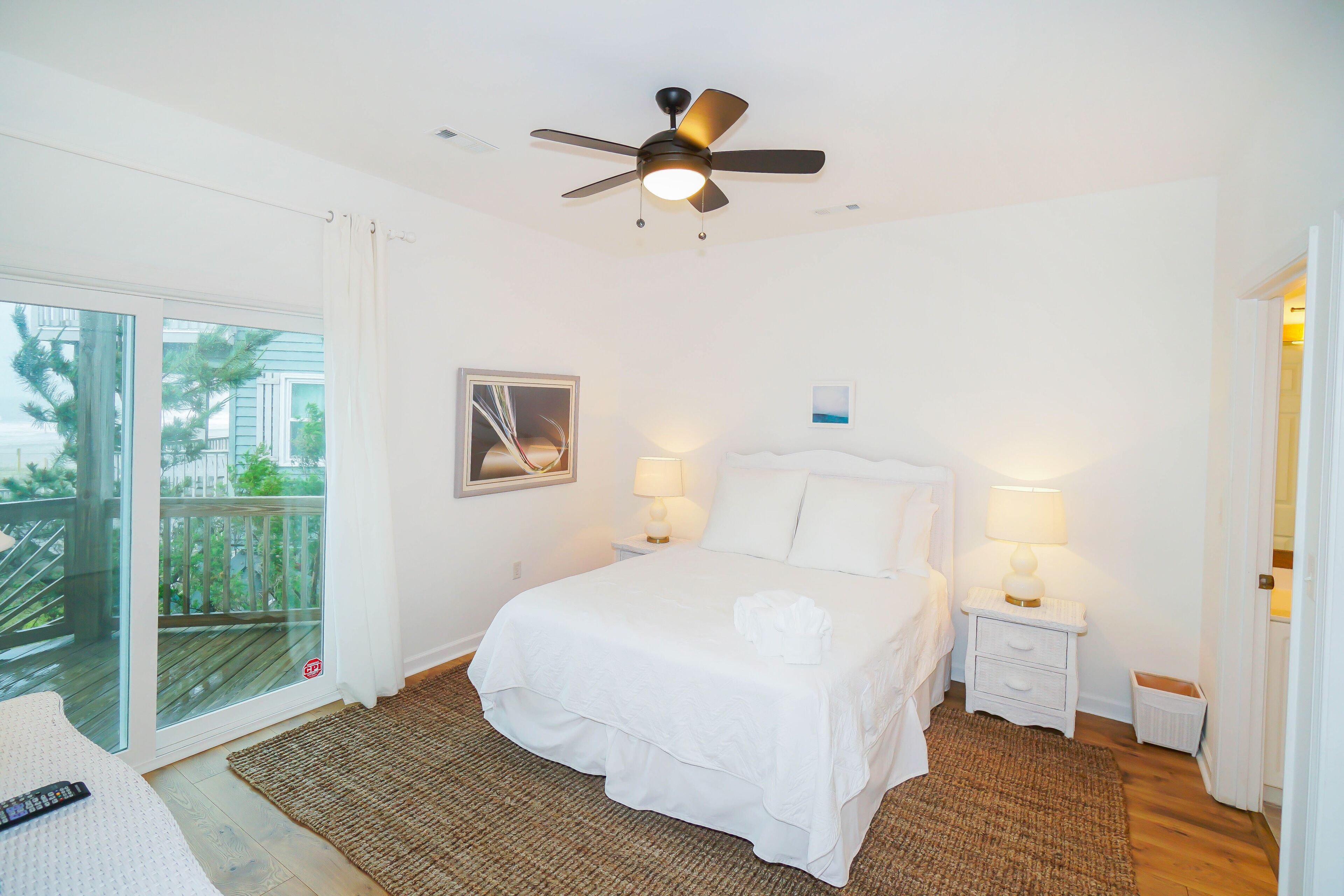 A hotel room with a bed, two bedside tables with lamps, a ceiling fan, a large window with a view of a balcony, and a framed picture on the wall.