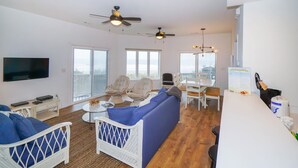 A spacious living area with a large sofa, two armchairs, a dining table, and a TV mounted on the wall.