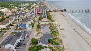 An aerial view of a coastal town with a beach, a pier, and a hotel with multiple floors and balconies.