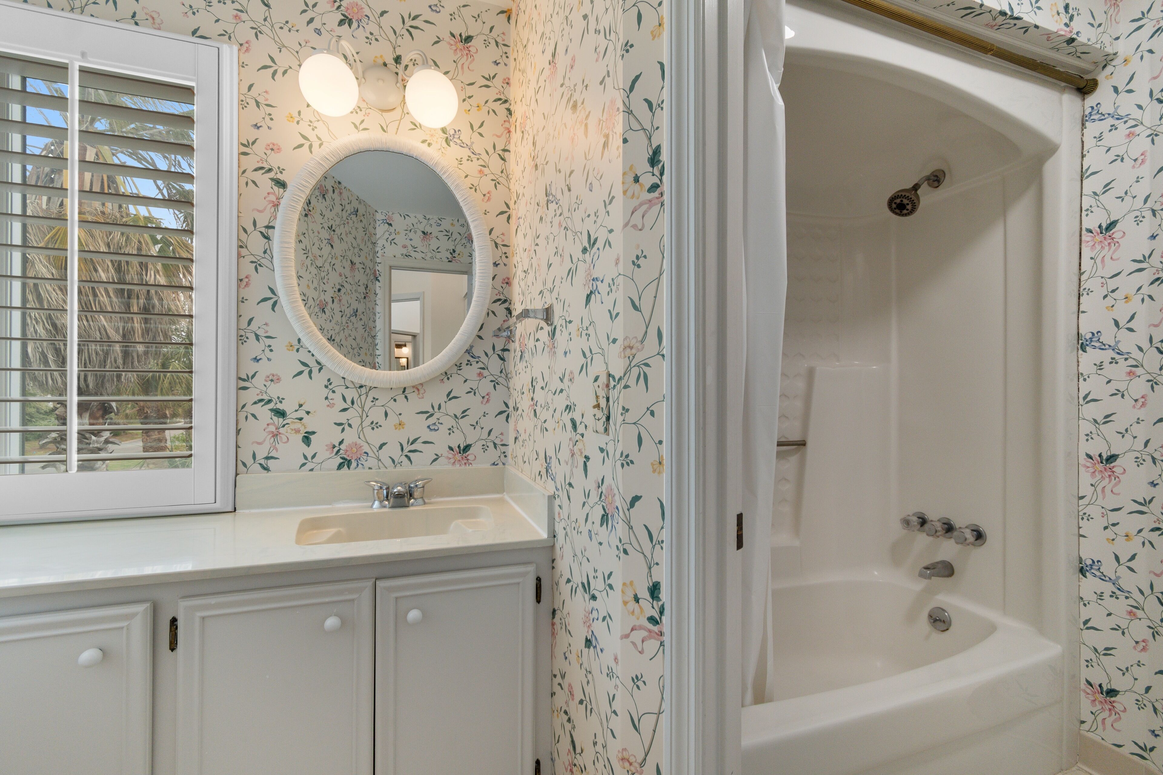 A bathroom with a floral wallpaper, a round mirror, a sink, and a bathtub.