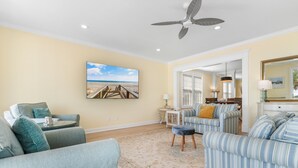 A living room with a large flat-screen TV, a ceiling fan, and a view of the beach through the window. - 7BR Beach House with Private Pool and Beach Access (Garden City Beach)