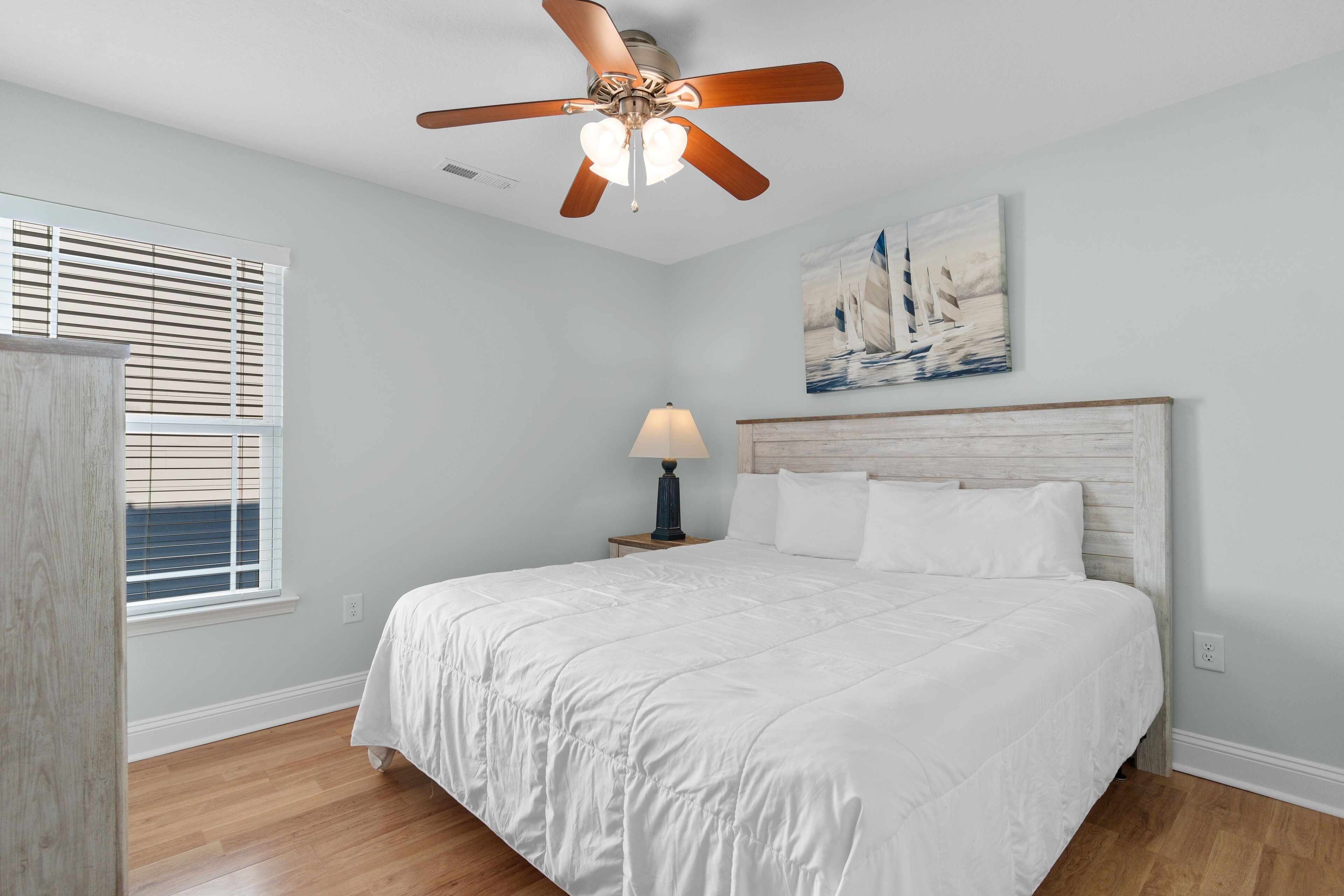 A bedroom with a bed, a ceiling fan, a nightstand with a lamp, a window with blinds, and a painting on the wall.