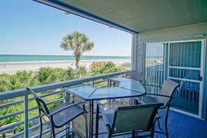 Ein Balkon mit Glastisch und Stühlen, mit Blick auf einen Strand mit Palmen und einem klaren Himmel.