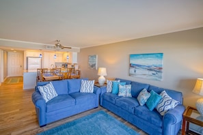 Ein Wohnzimmer mit einem blauen Ecksofa, einem Essbereich mit Tisch und Stühlen sowie einer Küche im Hintergrund.