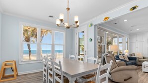 Un espace repas comprenant une table et des chaises, un lustre et une vue sur la plage grâce à de grandes fenêtres.