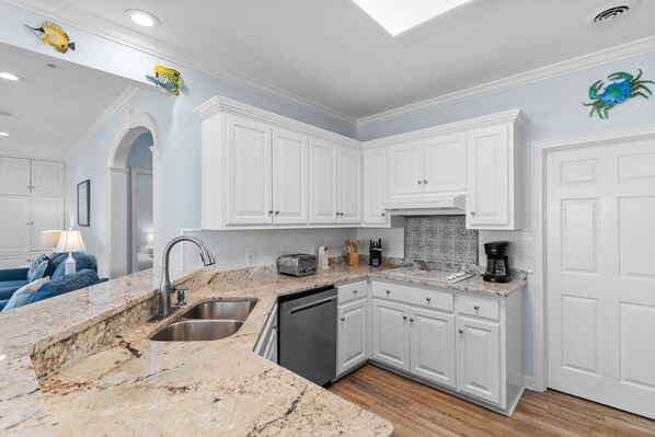 A kitchen with white cabinets, a granite countertop, a dishwasher, and a coffee maker. - 7BR Home w/ Beach Access & Stunning Ocean Views (Garden City)
