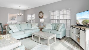 A living room with two light blue sofas, a wooden coffee table, a TV stand, and a large window with blinds. - 5BR Ocean-Side Home w/ Private Pool & Ocean Views Near Surfside Pier (Surfside Beach)