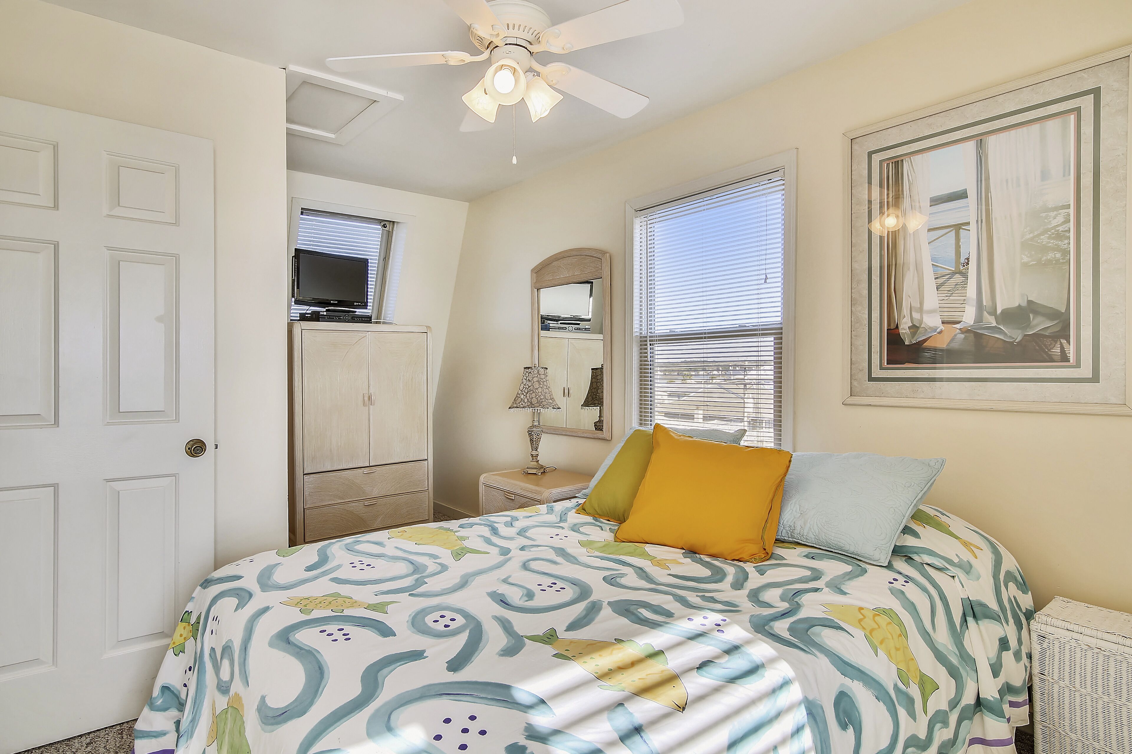A bedroom with a bed, a ceiling fan, a TV on a stand, a window with blinds, and a framed picture on the wall.