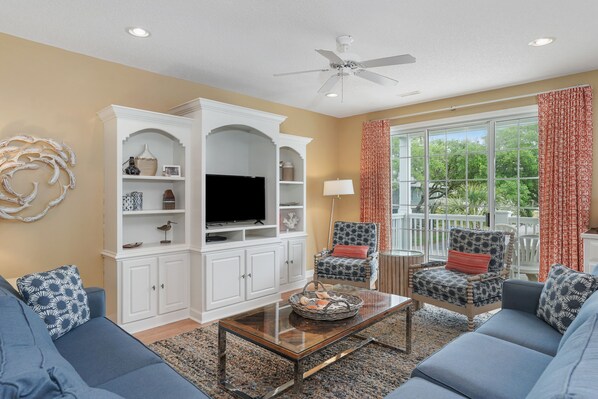A living room with a TV, white cabinets, blue sofas, and a large window with curtains.