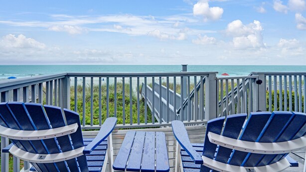 Une terrasse avec deux chaises Adirondack bleues et une table en bois, donnant sur une plage, elle-même bordée d’une rambarde et d’un escalier menant à l’eau.