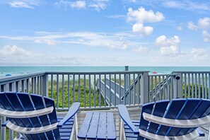 Een terras met twee blauwe Adirondack-stoelen en een houten tafel, met uitzicht op een strand met een balustrade en een trap naar het water.