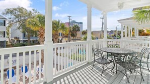 A covered outdoor area with a white railing, a round table with chairs, and a view of surrounding buildings and trees. - Walk to Beach Surfside Beach Home w/ Private Pool & Linens (Surfside Beach)