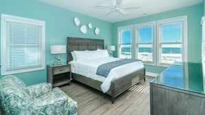 Una camera da letto con un letto, comodini, una sedia e vista sulla spiaggia attraverso ampie finestre.
