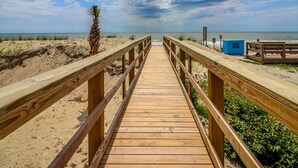 Una passerella di legno che conduce a una spiaggia, con una palma e una struttura blu sulla destra.