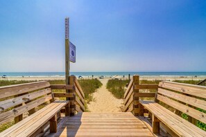 A wooden boardwalk leading to a beach with a signpost and a clear view of the ocean.