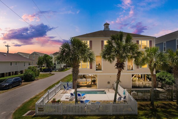 A two-story house with a swimming pool, palm trees, and a white picket fence.