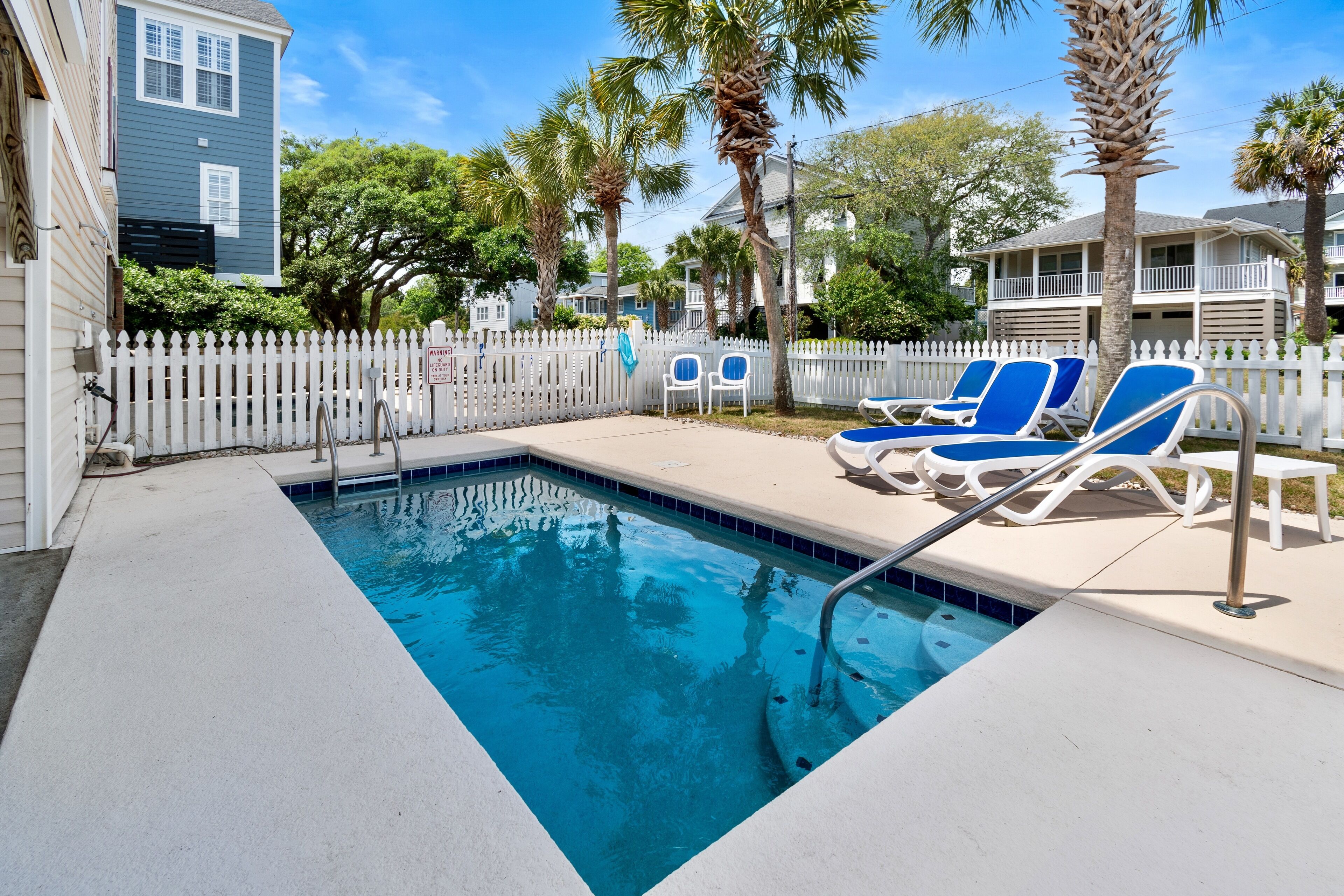A swimming pool with blue water, surrounded by a white picket fence, and blue lounge chairs.
