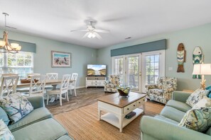A spacious living room with a dining area, a ceiling fan, and a large window with blinds.