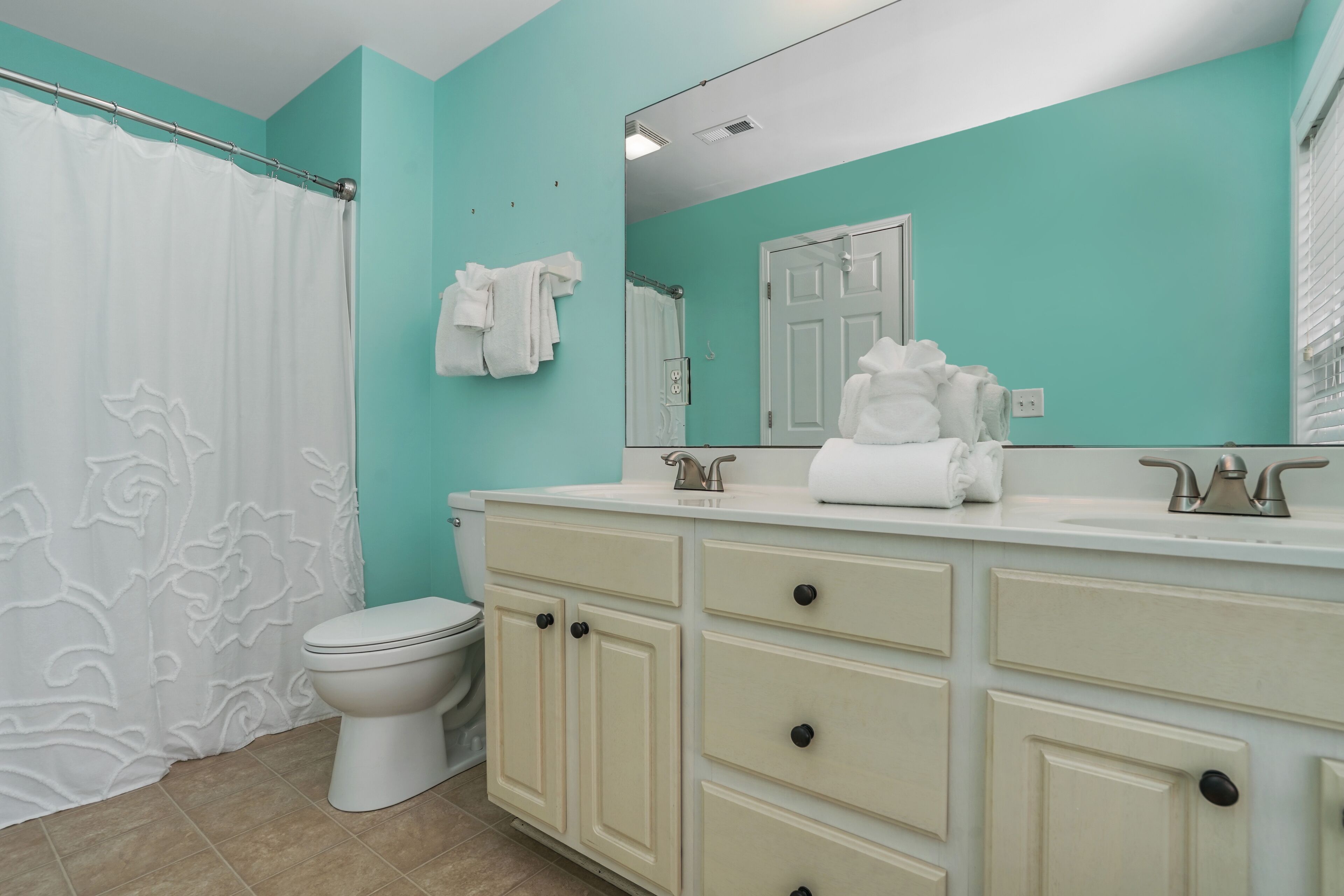 A bathroom with a white toilet, a white shower curtain, a white sink with a mirror above it, and a teal wall.