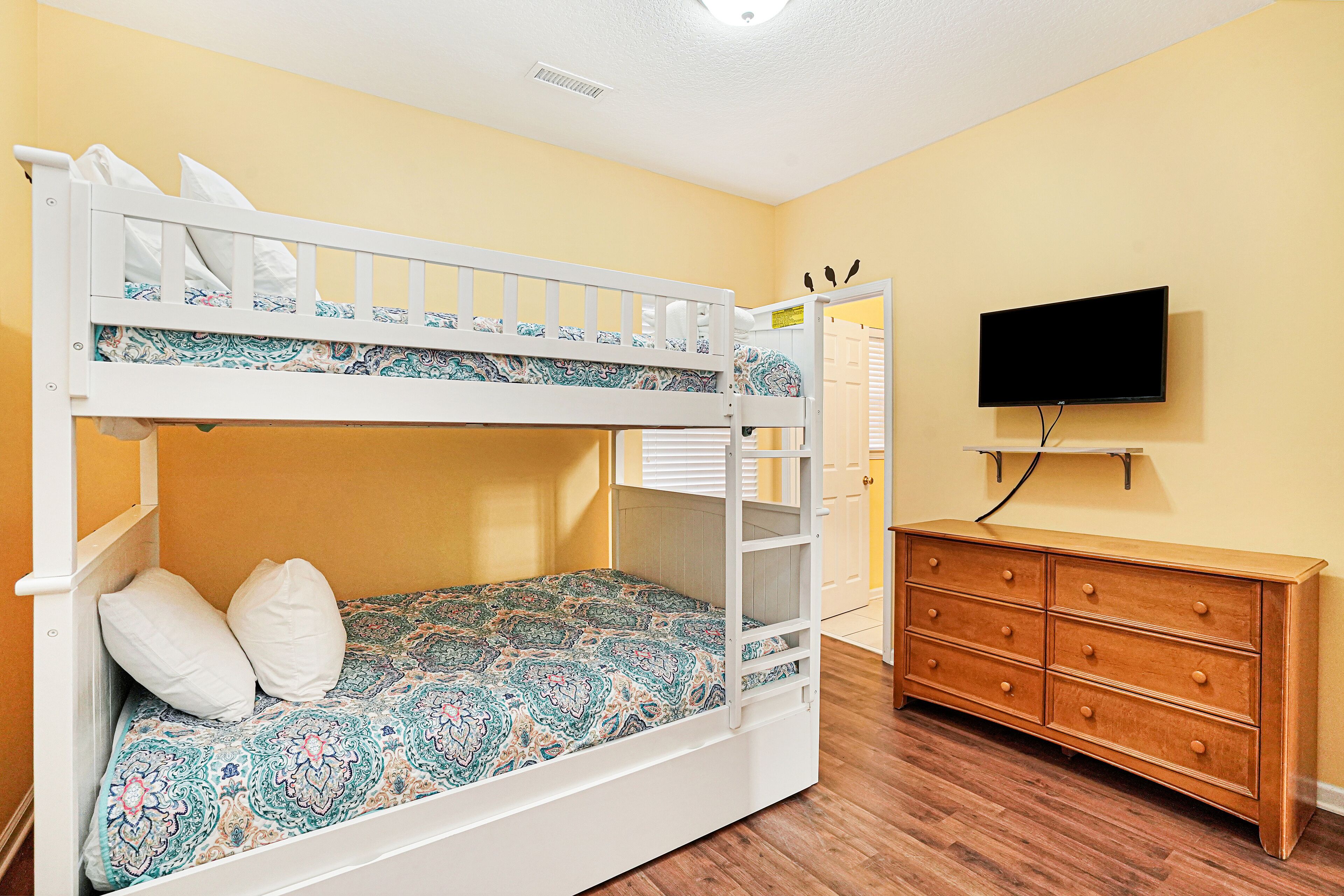 A bunk bed with a patterned bedspread, a wooden dresser, and a flat-screen TV mounted on the wall.