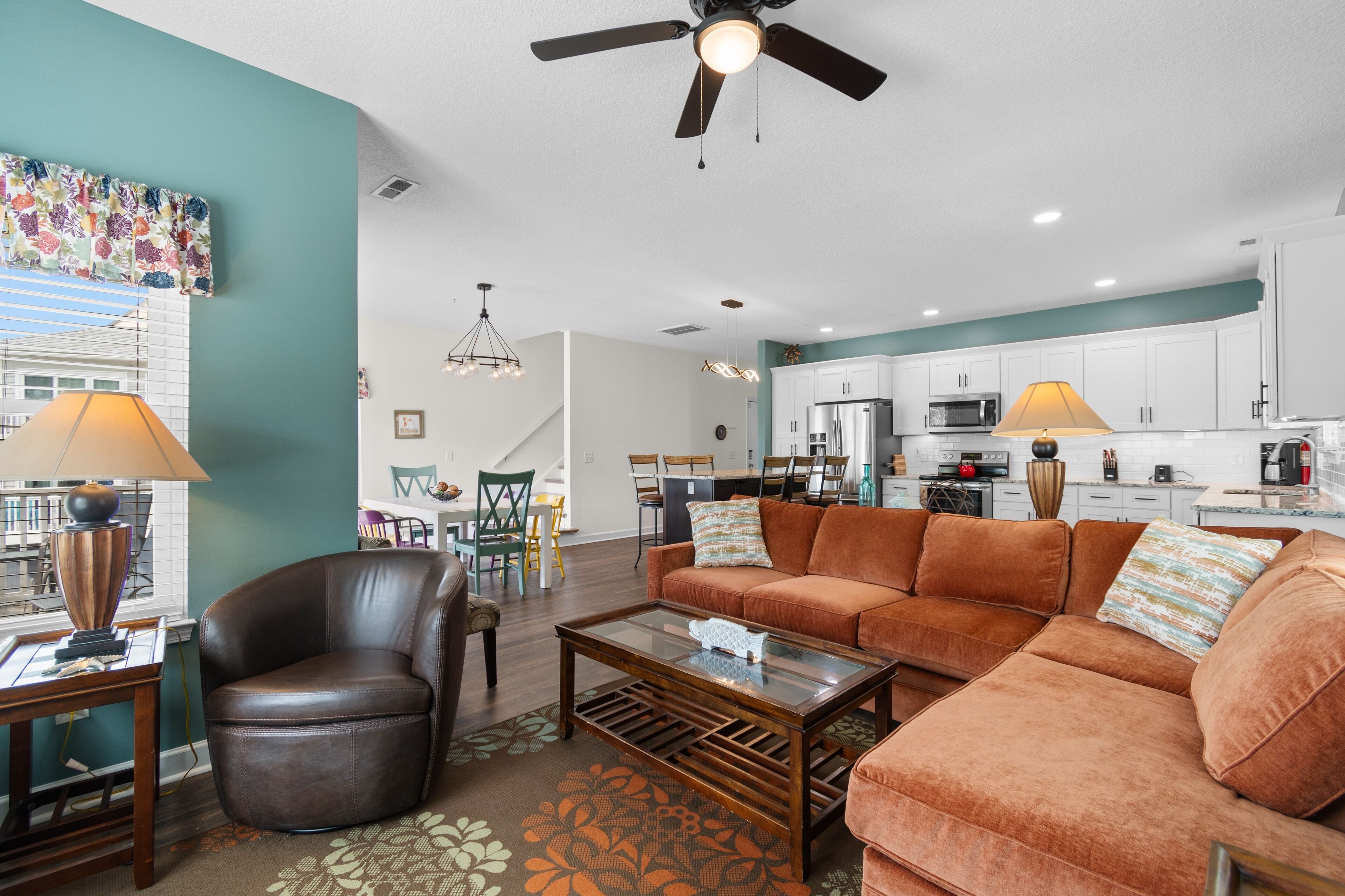 A spacious living room with a brown leather chair, a glass coffee table, and a brown sofa. There is a dining area with a table and chairs, and a kitchen with white cabinets and appliances.
