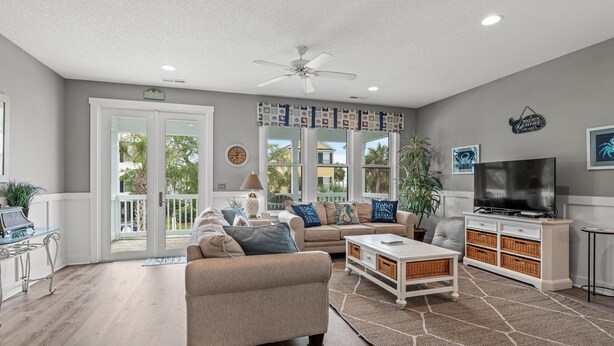 A spacious living room with a sofa, coffee table, and TV stand, featuring a large window and a ceiling fan. - Walk to Beach Surfside House w/ Pool & 4 Bedrooms (Surfside Beach)