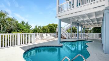A swimming pool with a white deck, a covered upper level with a balcony, and a white fence surrounding the area.