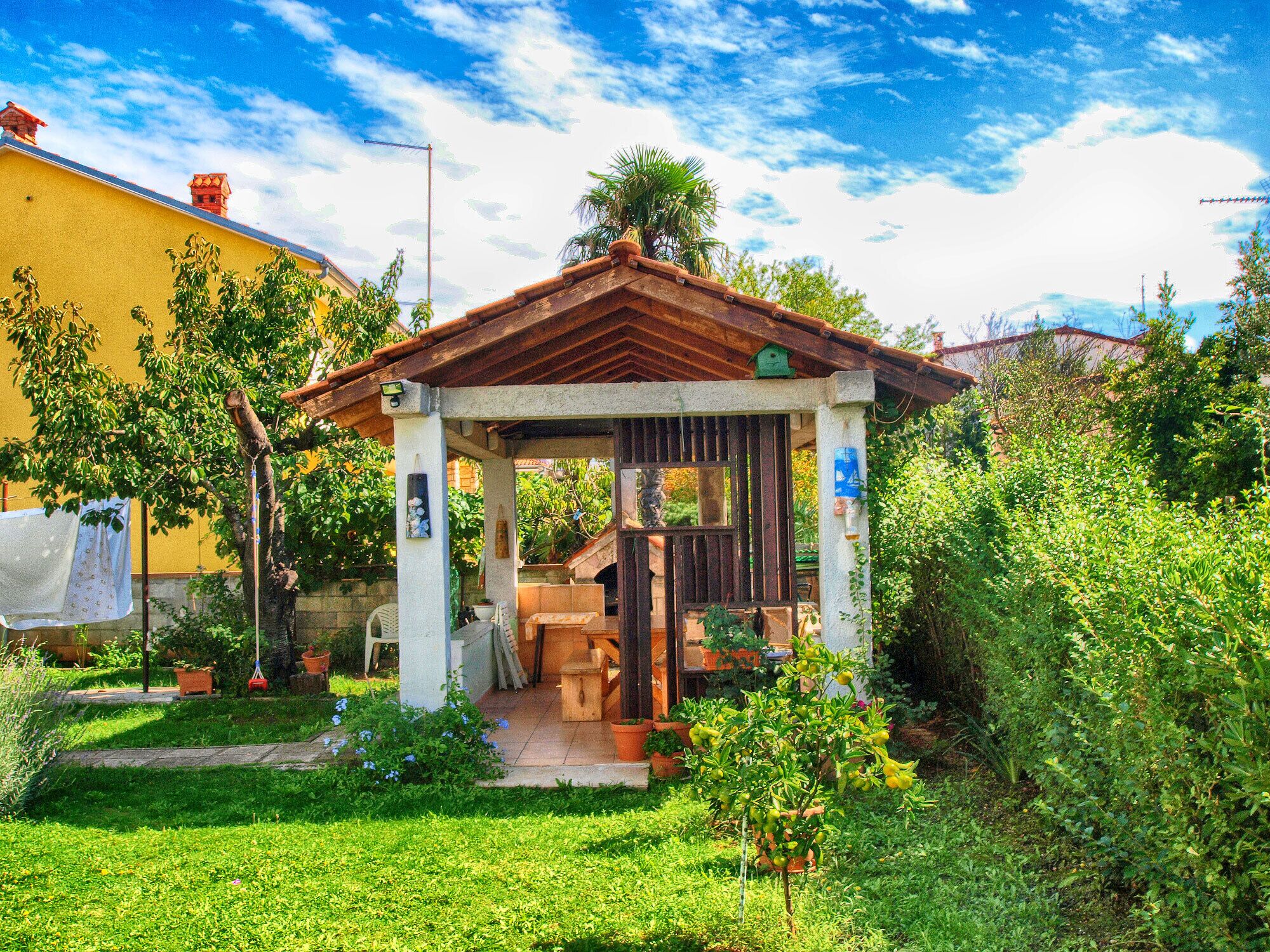 A small, rustic gazebo with a tiled roof, surrounded by lush greenery and a yellow building in the background.