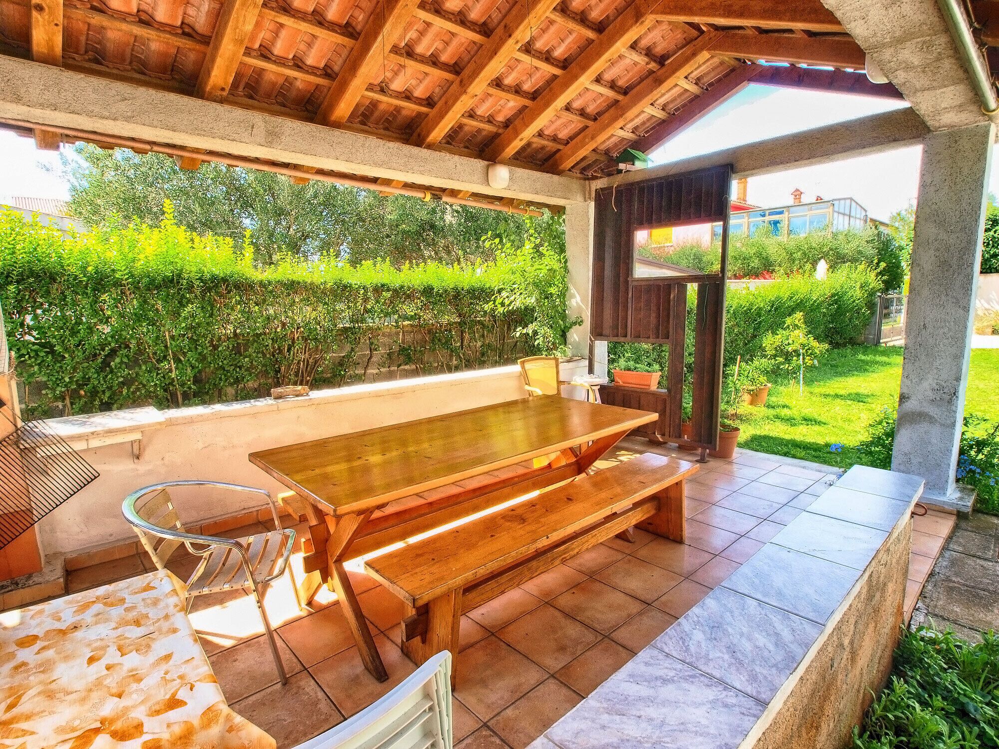 Une terrasse extérieure avec une table et des bancs en bois, entourée de verdure et dotée d’un sol carrelé.