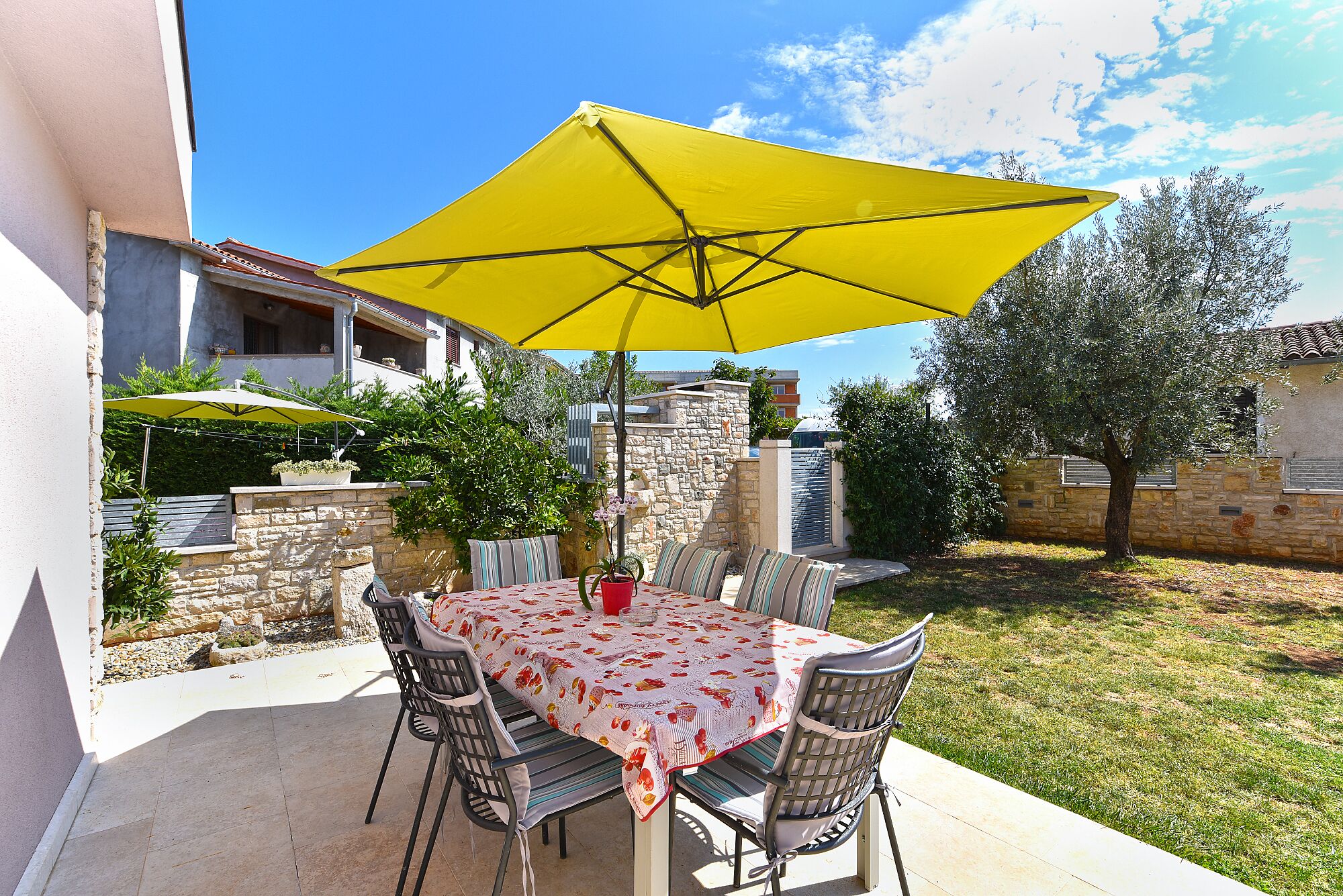 An outdoor patio with a table and chairs, a yellow umbrella, and a stone wall in the background.