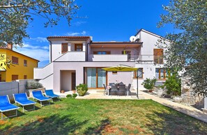 Una casa de dos pisos con un balcón, un patio con mesa y sillas, y un jardín con sillones reclinables azules.