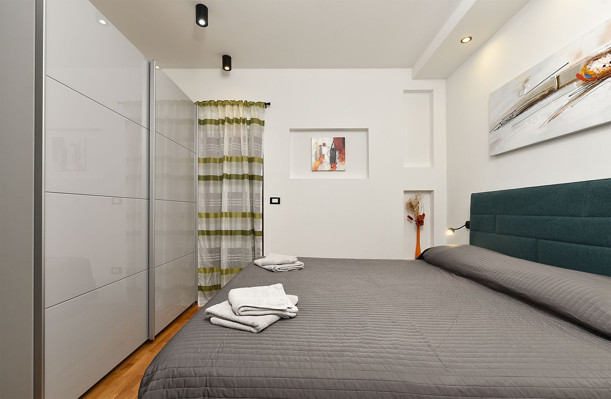 A modern bedroom with a large bed, a wardrobe, a shower curtain, a painting, and a vase with dried flowers.