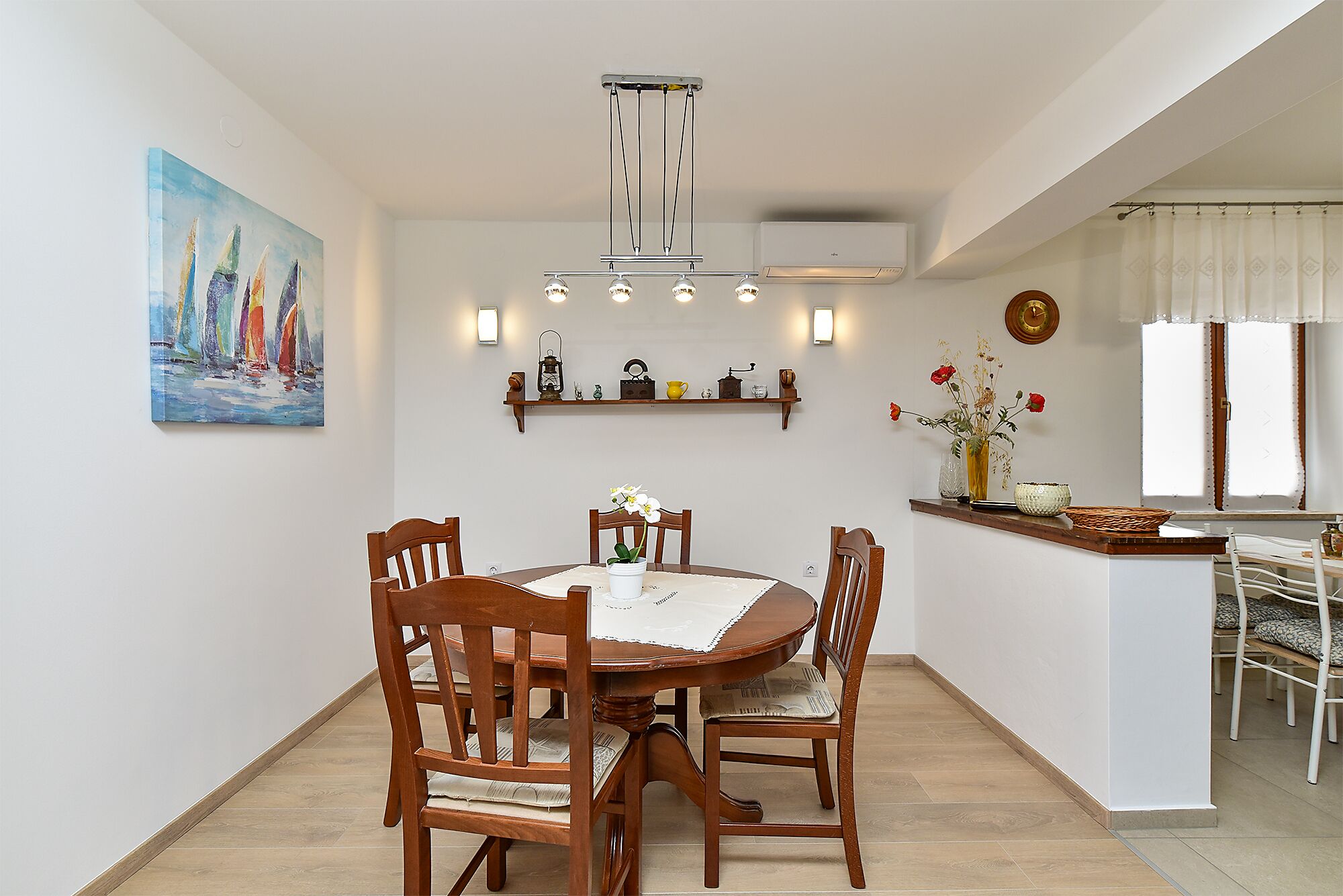 Un coin repas avec une table ronde en bois, quatre chaises, un tableau au mur, une étagère avec des objets décoratifs et une fenêtre avec des rideaux.