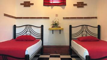 Een kamer met twee bedden, een geruite vloer, een plafondventilator, een raam met rode gordijnen en een klein plankje met een plant.