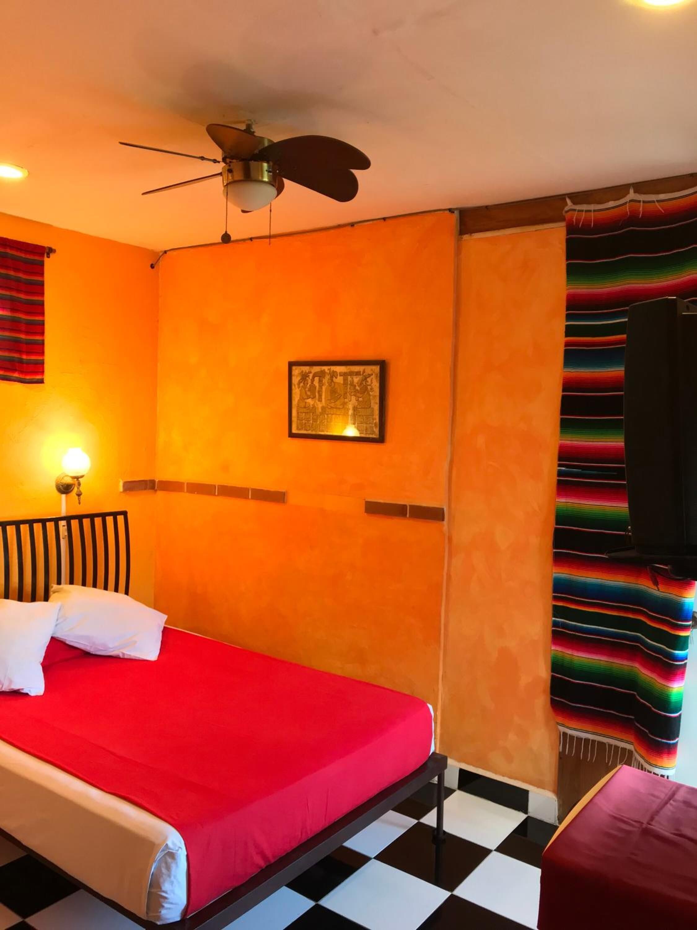 Ein Hotelzimmer mit roter Bettdecke, Deckenventilator, schwarz-weiß kariertem Boden und farbenfrohem Vorhang.