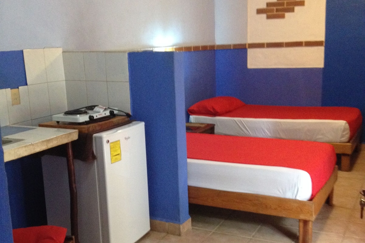 Ein kleines, kompaktes Zimmer mit Küchenzeile, einem Einzelbett und einem Sessel.