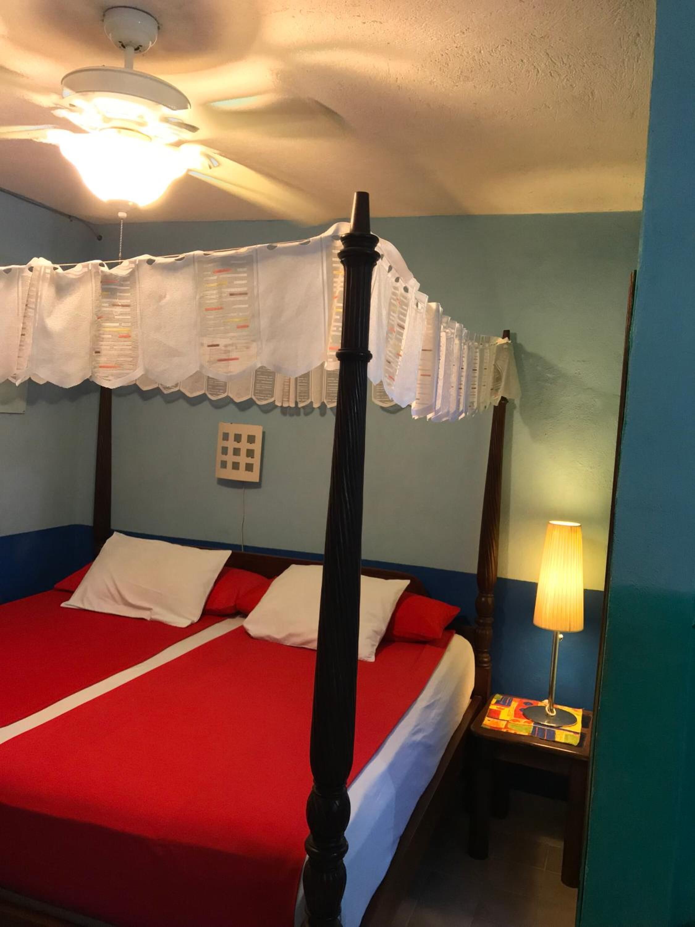 Una cama con dosel con un edredón rojo y blanco, un ventilador de techo y una mesita de noche con lámpara.
