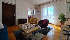 A living room with a wooden coffee table, a blue sofa, a yellow armchair, and a dark armchair. There is a potted plant on a side table and a window with curtains.