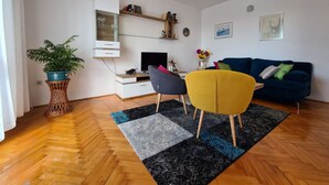 A modern living room with a wooden floor, a sofa, a chair, a TV, a small table, and a bookshelf.