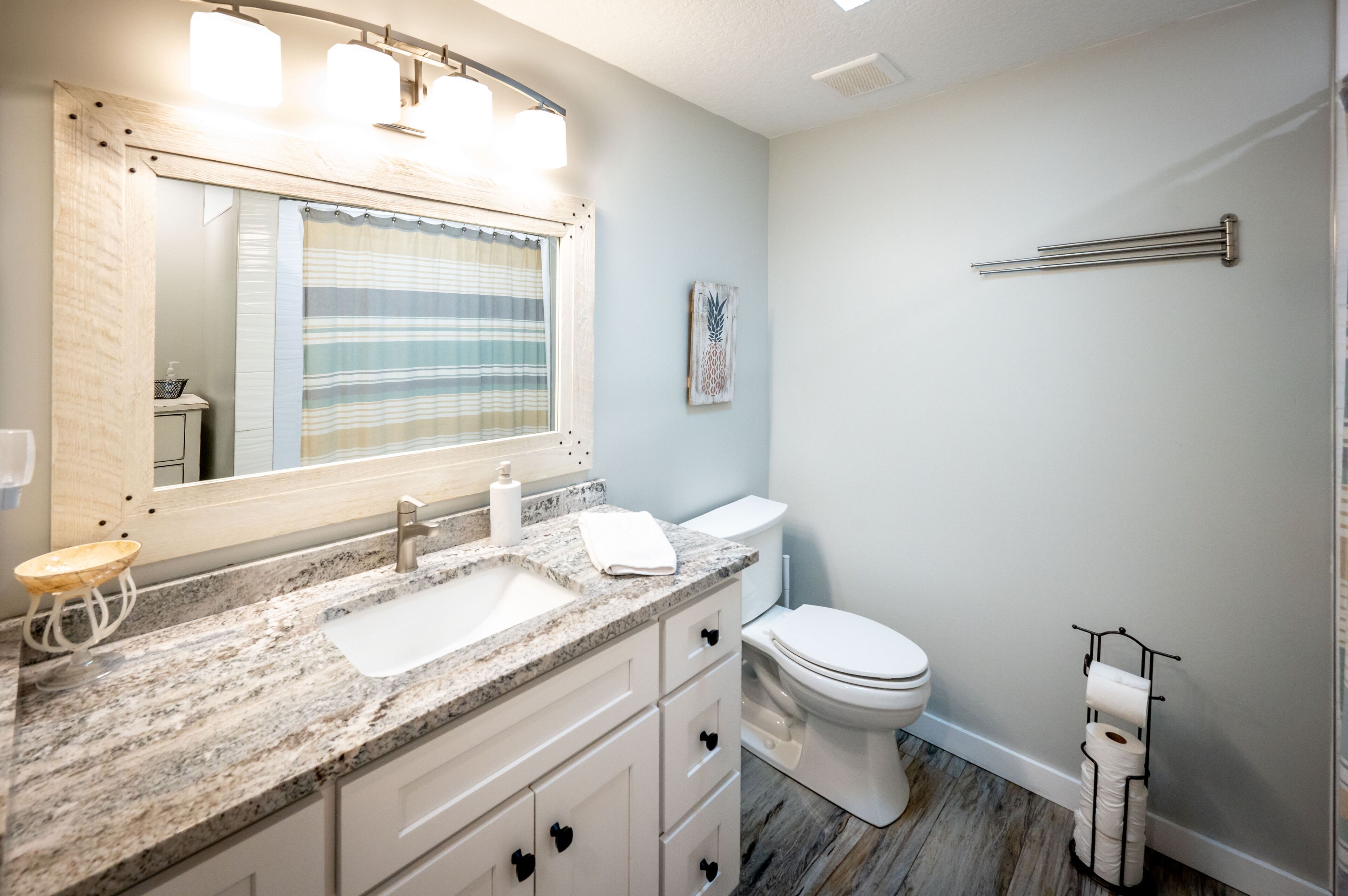 Une salle de bain avec un comptoir en granit, un lavabo blanc, un miroir avec éclairage intégré, des toilettes et un porte-serviettes.