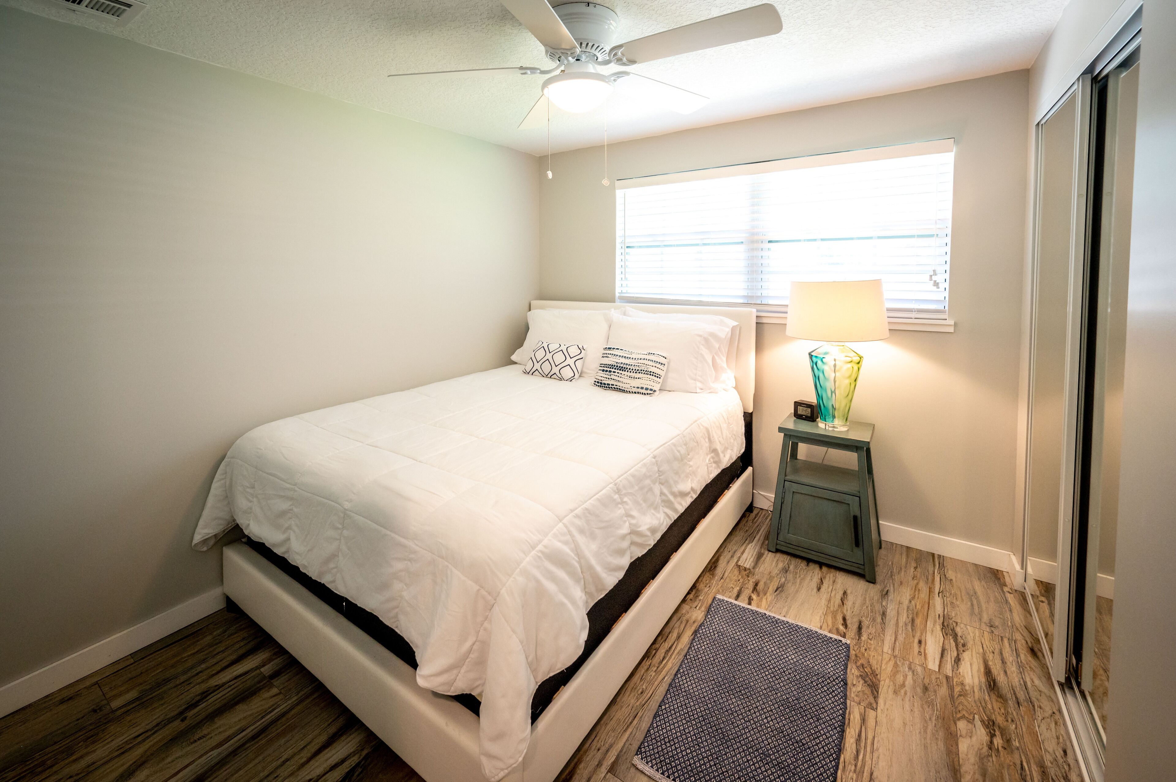 A bedroom with a bed, a ceiling fan, a window with blinds, and a nightstand with a lamp.