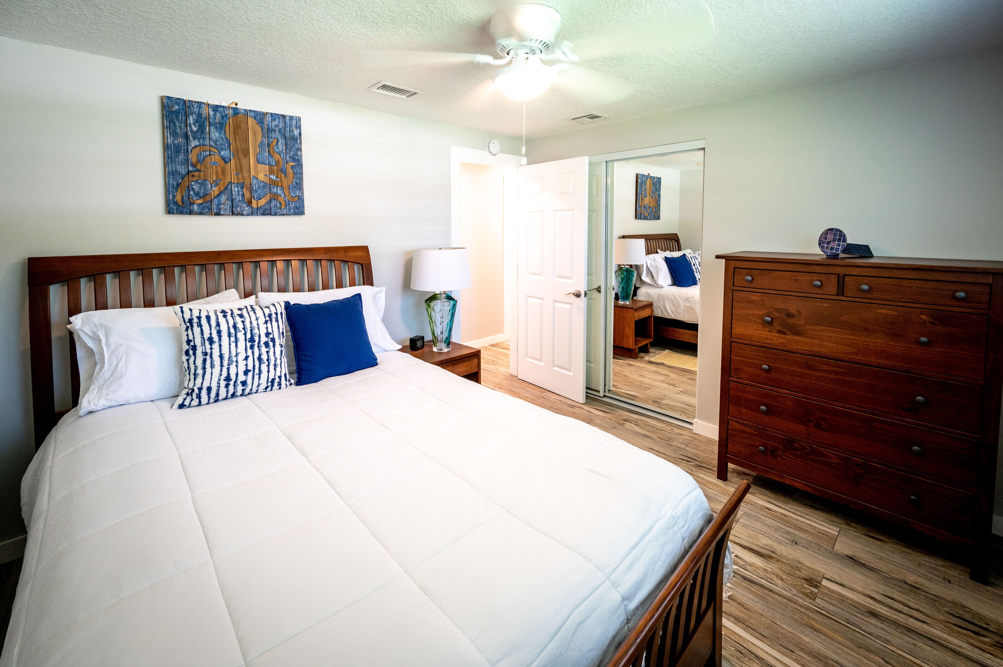 A bedroom with a wooden bed, a nightstand with a lamp, a dresser, and a ceiling fan.