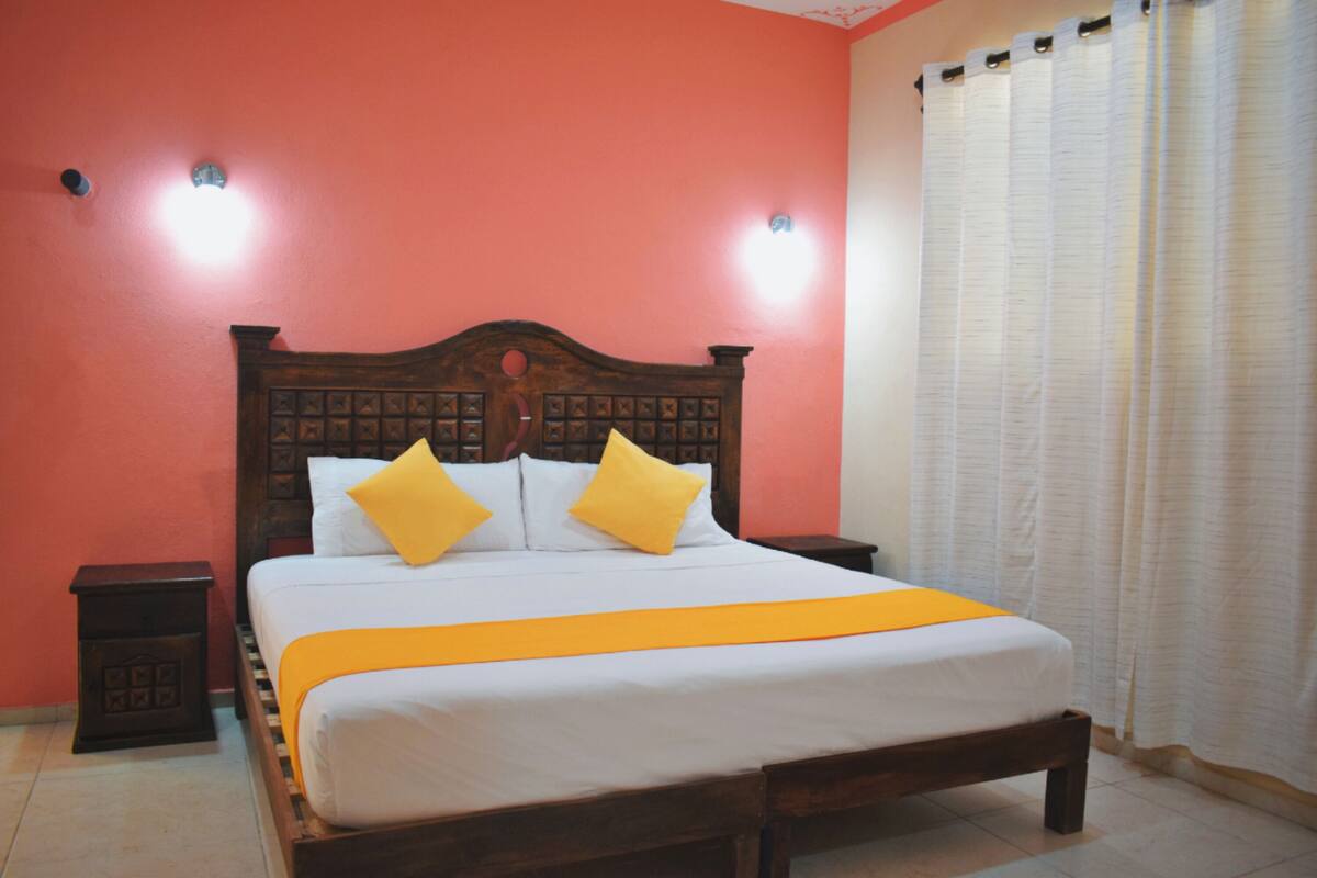 A bed with a wooden headboard, two pillows, and an orange bedspread in a room with red walls and a window with curtains.