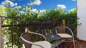 Een balkon met twee rieten stoelen en een klein rond tafeltje, omgeven door weelderige groene beplanting en een helderblauwe lucht.