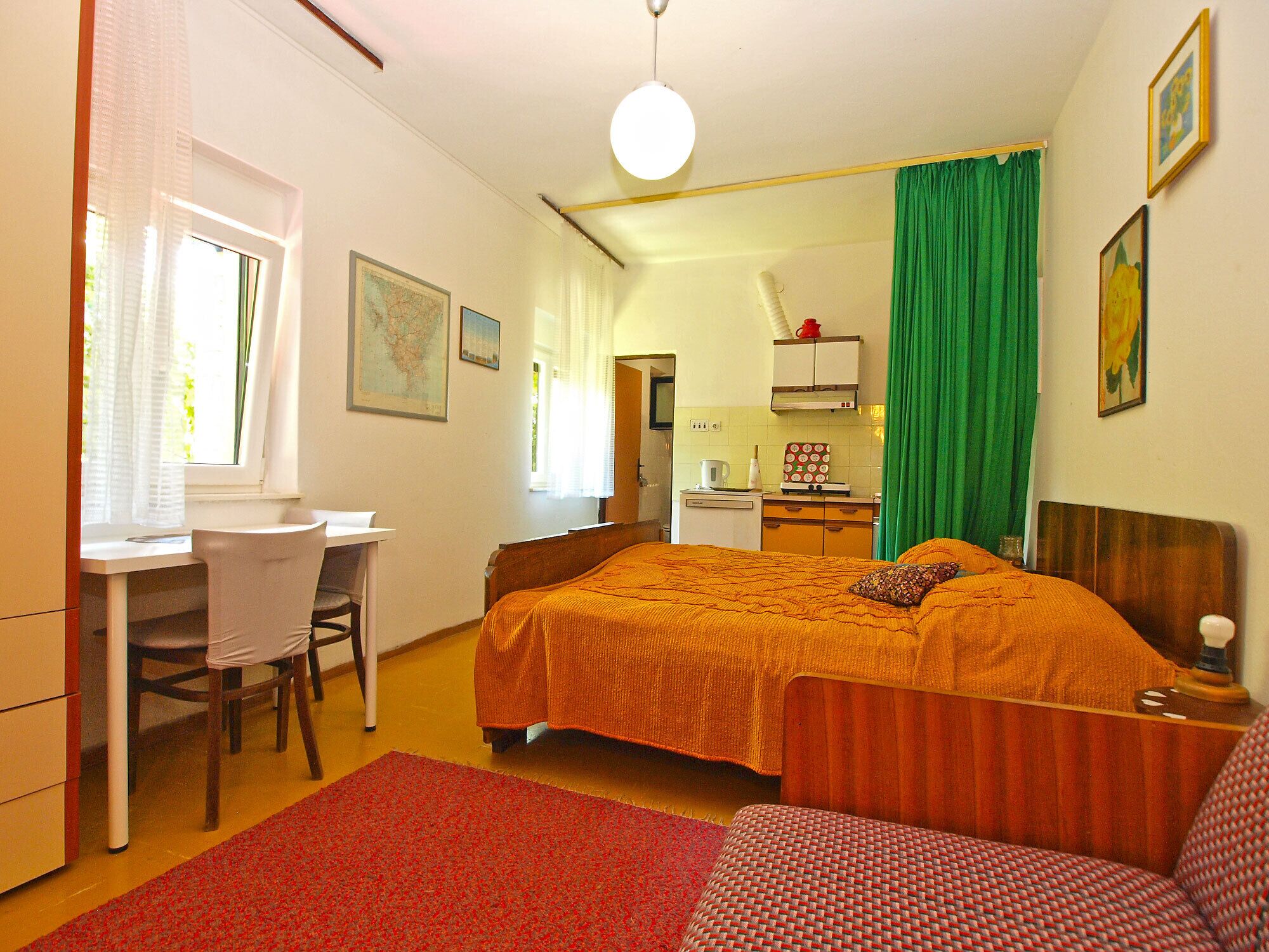 A bedroom with a bed, a desk, a chair, a kitchenette, and a bathroom.