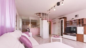A modern living room with a white sofa, a small table, and a TV. There is a staircase leading to an upper level.